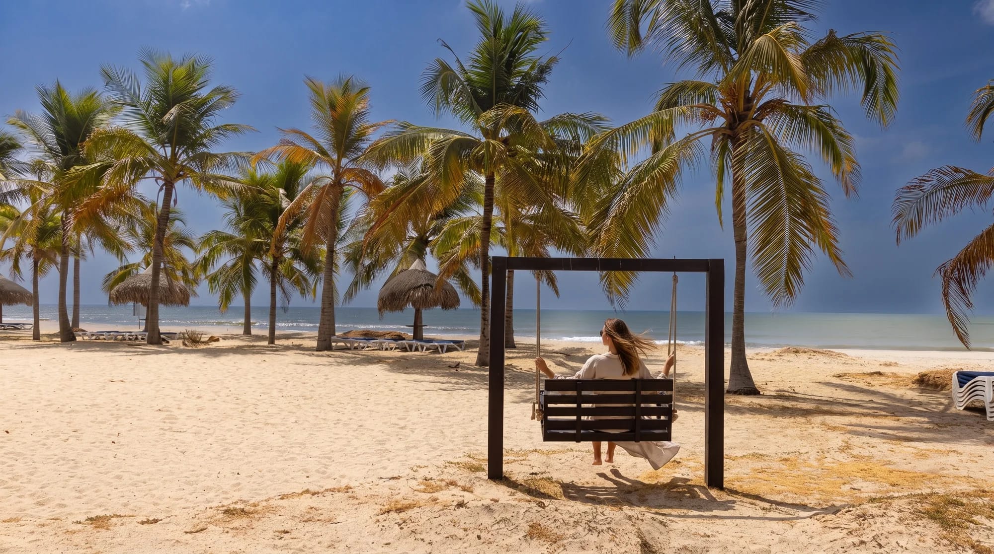 A woman sitting on a swing facing the sea, among palm trees